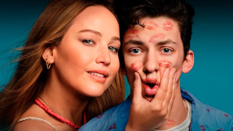 Image of a young woman scrunching a young man's face, which is peppered with lipstick lip-imprints, from the movie No Hard Feelings.